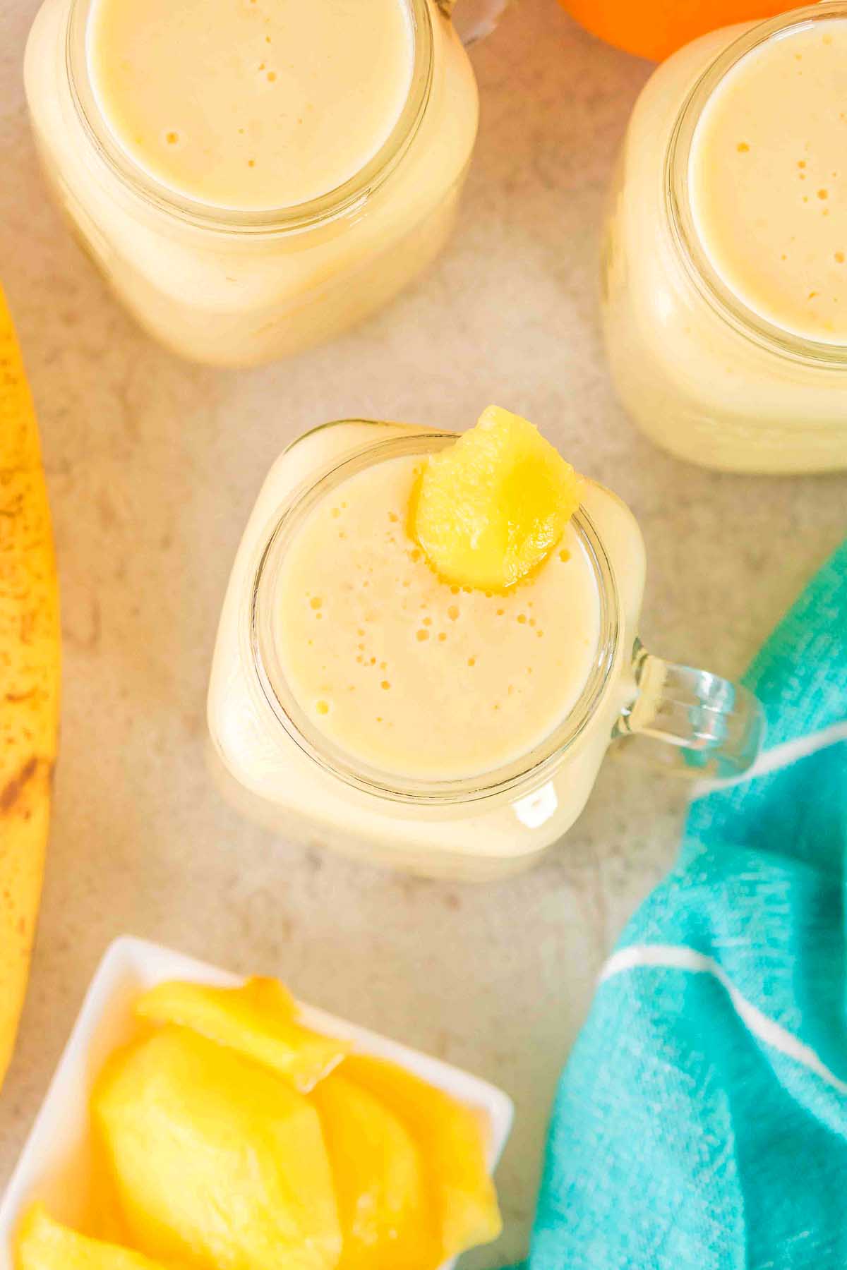 Three jars of creamy yellow Pineapple Mango Smoothie, one topped with a pineapple chunk, sit next to a teal cloth and fresh fruit pieces.