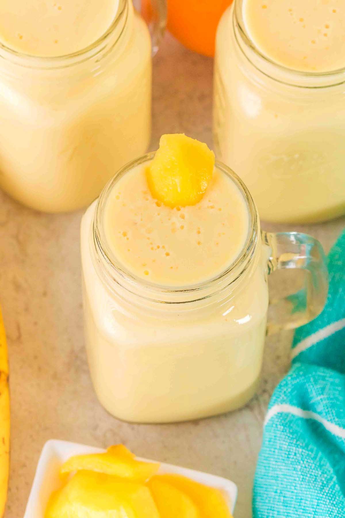 A glass jar of creamy yellow Pineapple Mango Smoothie, topped with a mango piece, surrounded by fruit and a blue cloth.