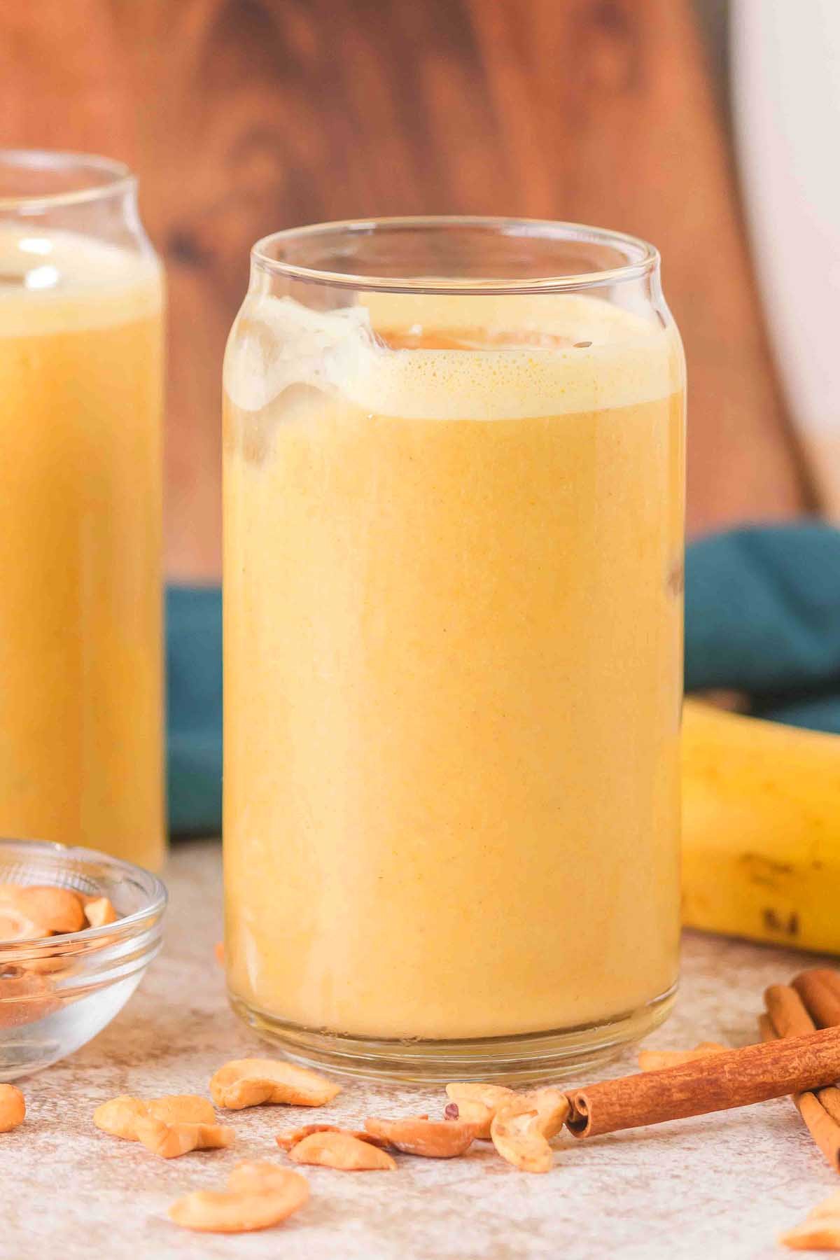 A glass of creamy Banana Cashew Smoothie with ice, surrounded by cashews, a cinnamon stick, and a fresh banana.
