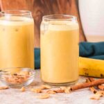 Two glasses of Banana Cashew Smoothie with ice, surrounded by a fresh banana, cashews, and cinnamon sticks on a table.