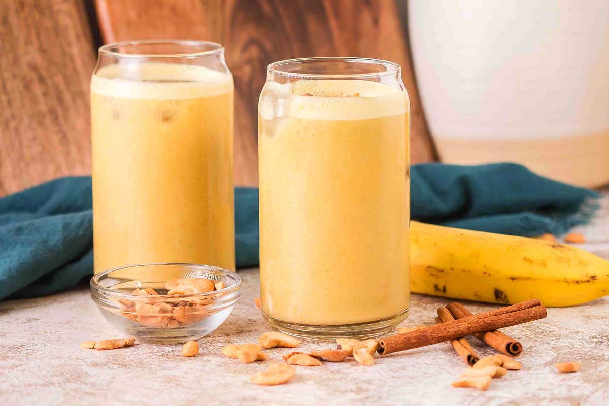 Two glasses of Banana Cashew Smoothie with ice, surrounded by a fresh banana, cashews, and cinnamon sticks on a table.
