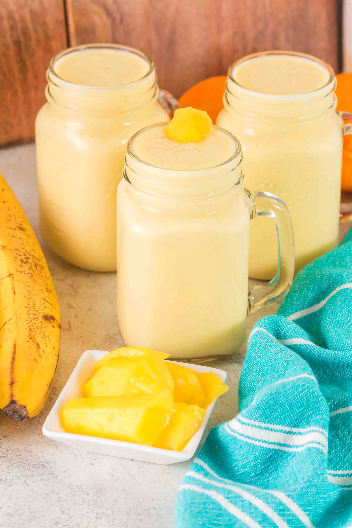 Three glass mugs of Pineapple Mango Smoothie with mango chunks, a banana, and a teal-striped cloth on the table.