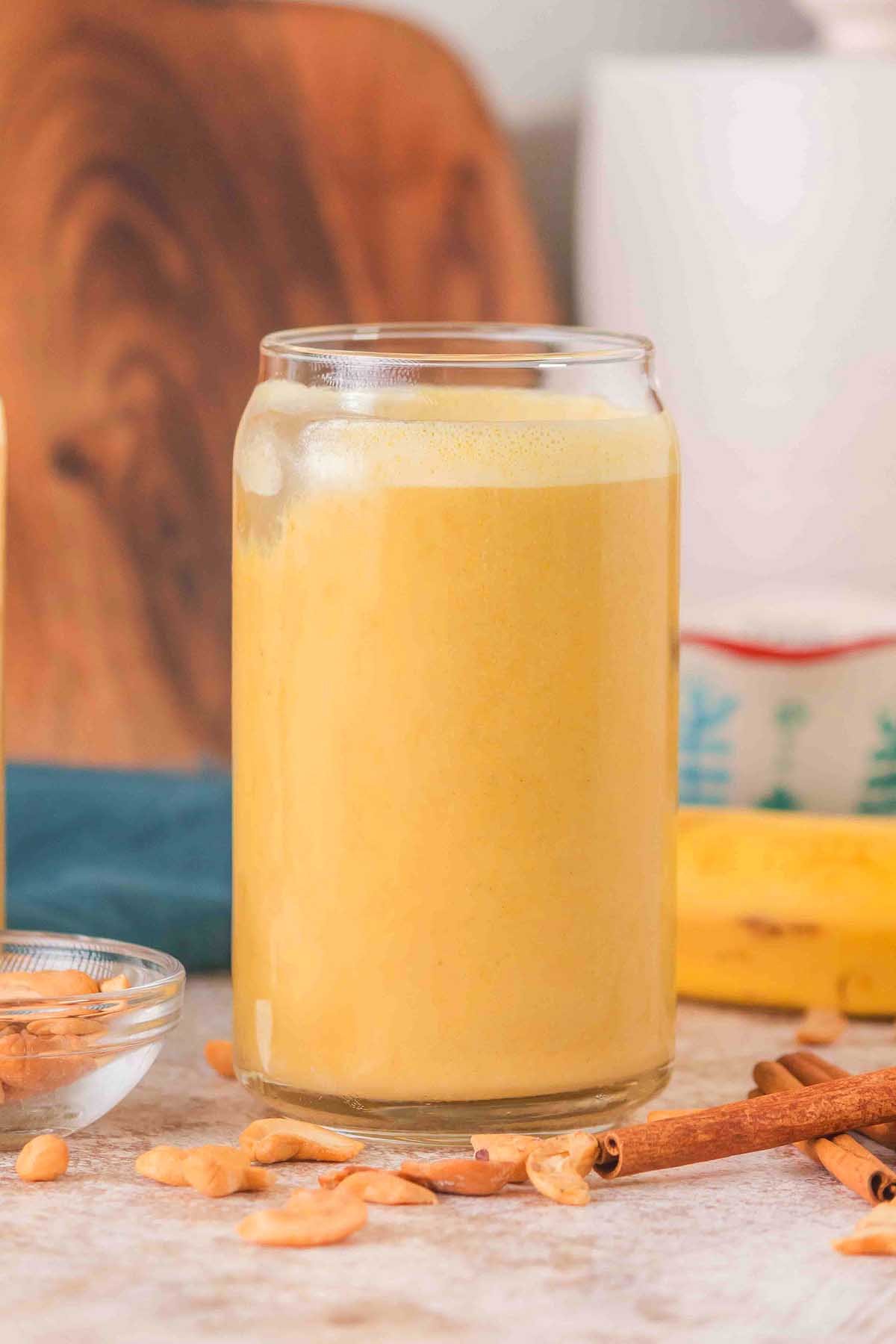 A glass of creamy yellow Banana Cashew Smoothie with peanuts, cinnamon sticks, and a banana in the background.
