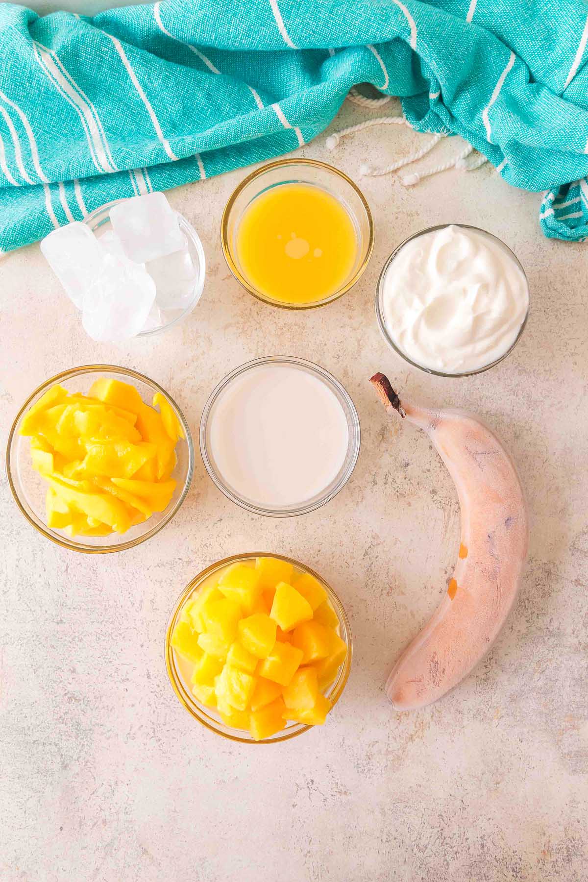 Six smoothie ingredients—ice cubes, orange juice, yogurt, banana, milk, and chopped mango—on a light surface, perfect for crafting a refreshing Pineapple Mango Smoothie.