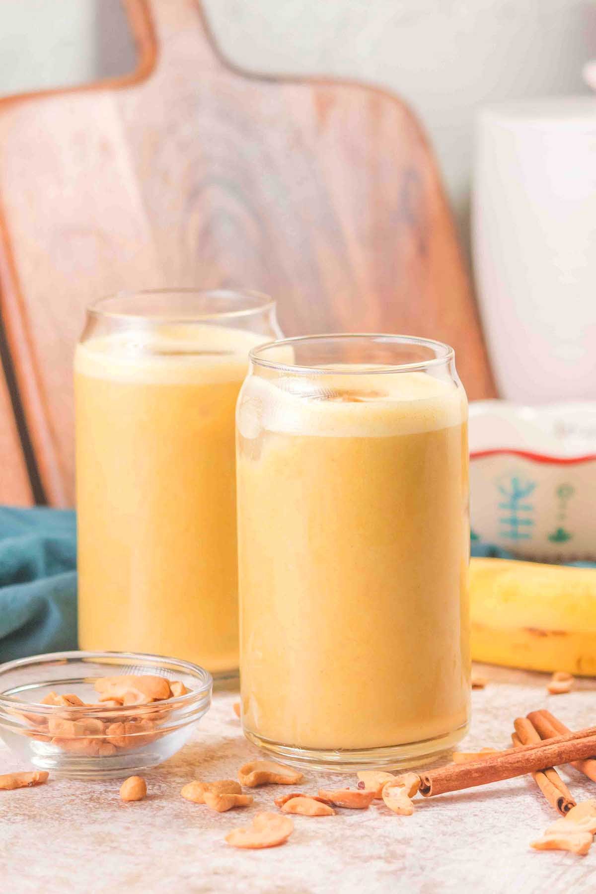 Two glasses of creamy Banana Cashew Smoothie on a table with peanuts and cinnamon sticks nearby.