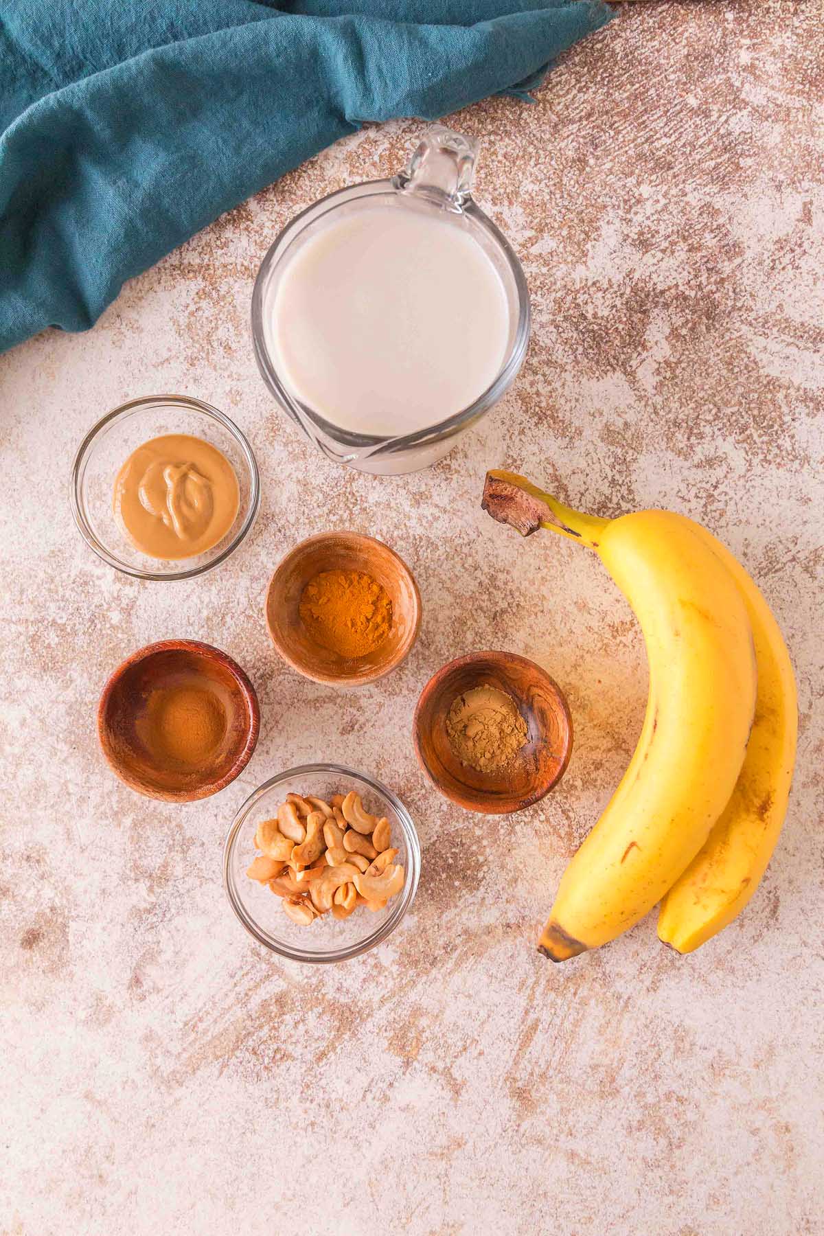 Overhead view of ingredients for a Banana Cashew Smoothie—bananas, cashews, peanut butter, spices, and a glass jug of milk on a textured surface.