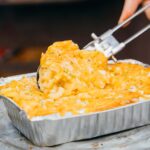 A person serving cheesy campfire mac and cheese from a foil tray with tongs.