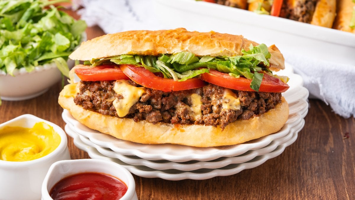 A Chopped Cheese Sandwich with ground beef, melted cheese, tomato slices, and lettuce on a plate, served with ketchup and mustard nearby.
