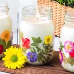 Three lit DIY pressed flower candle jars in glass, each decorated with colorful blooms, arranged gracefully on a wooden board.