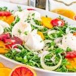 Fresh salad with arugula, fennel, oranges, blood oranges, and burrata on a white platter. Orange dressing on the side.