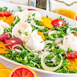 Fresh salad with arugula, fennel, oranges, blood oranges, and burrata on a white platter. Orange dressing on the side.