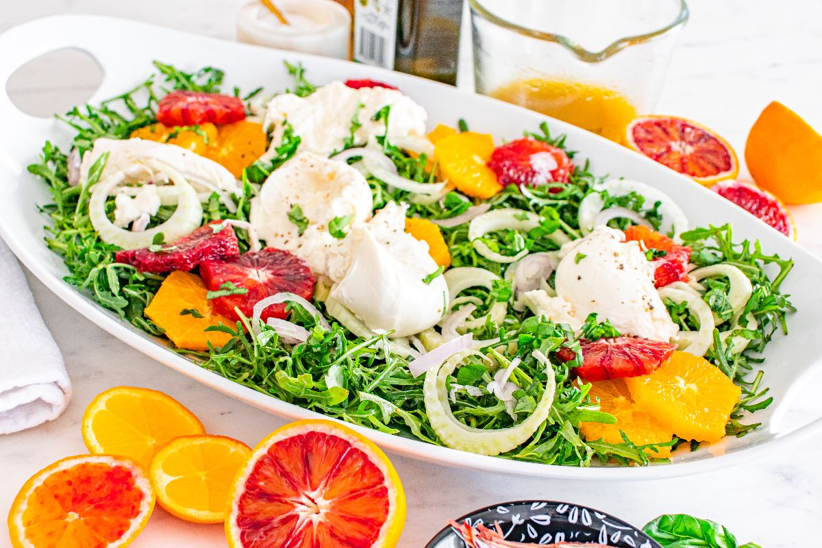 A fresh salad with greens, sliced citrus fruits, fennel, and dollops of cheese on a white platter.