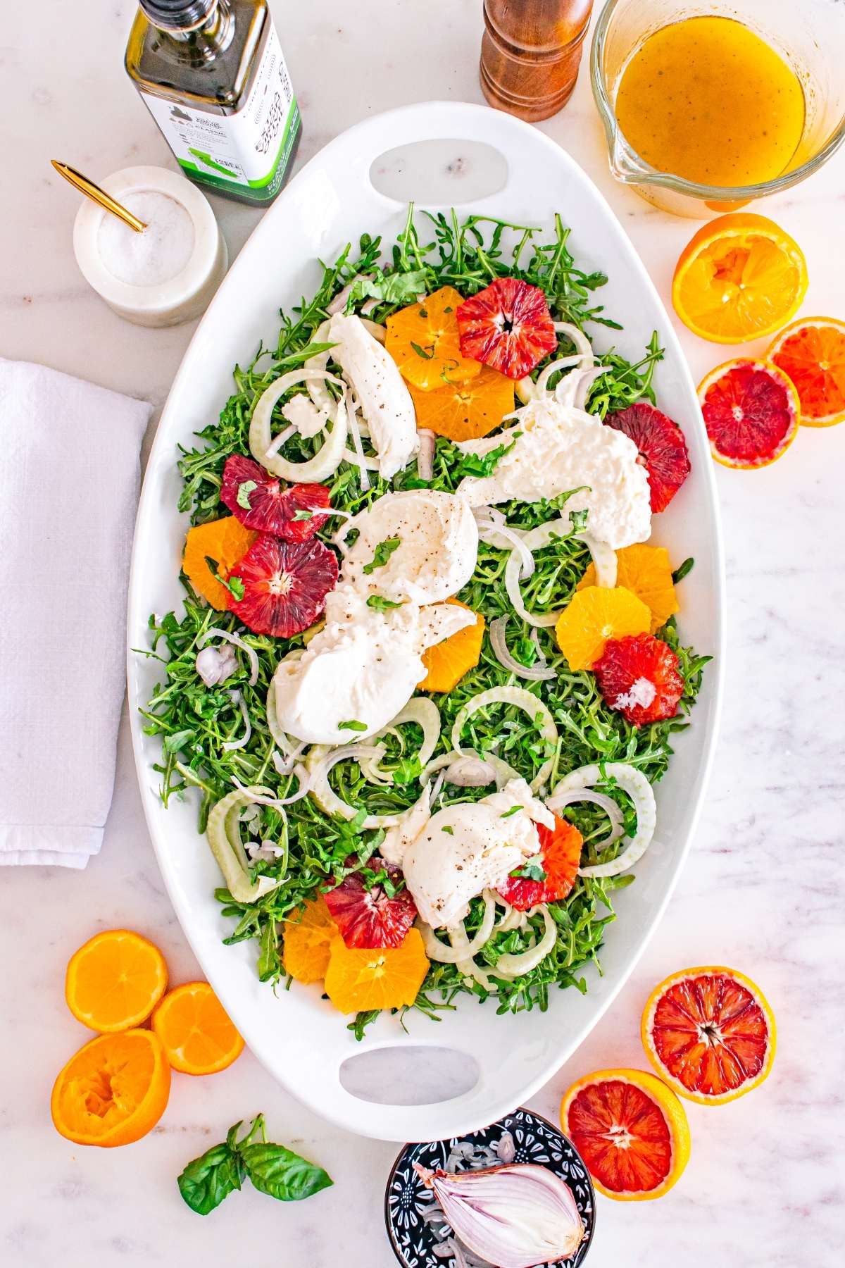 Salad with arugula, citrus slices, burrata, and fennel on a white platter, surrounded by dressing and ingredients.