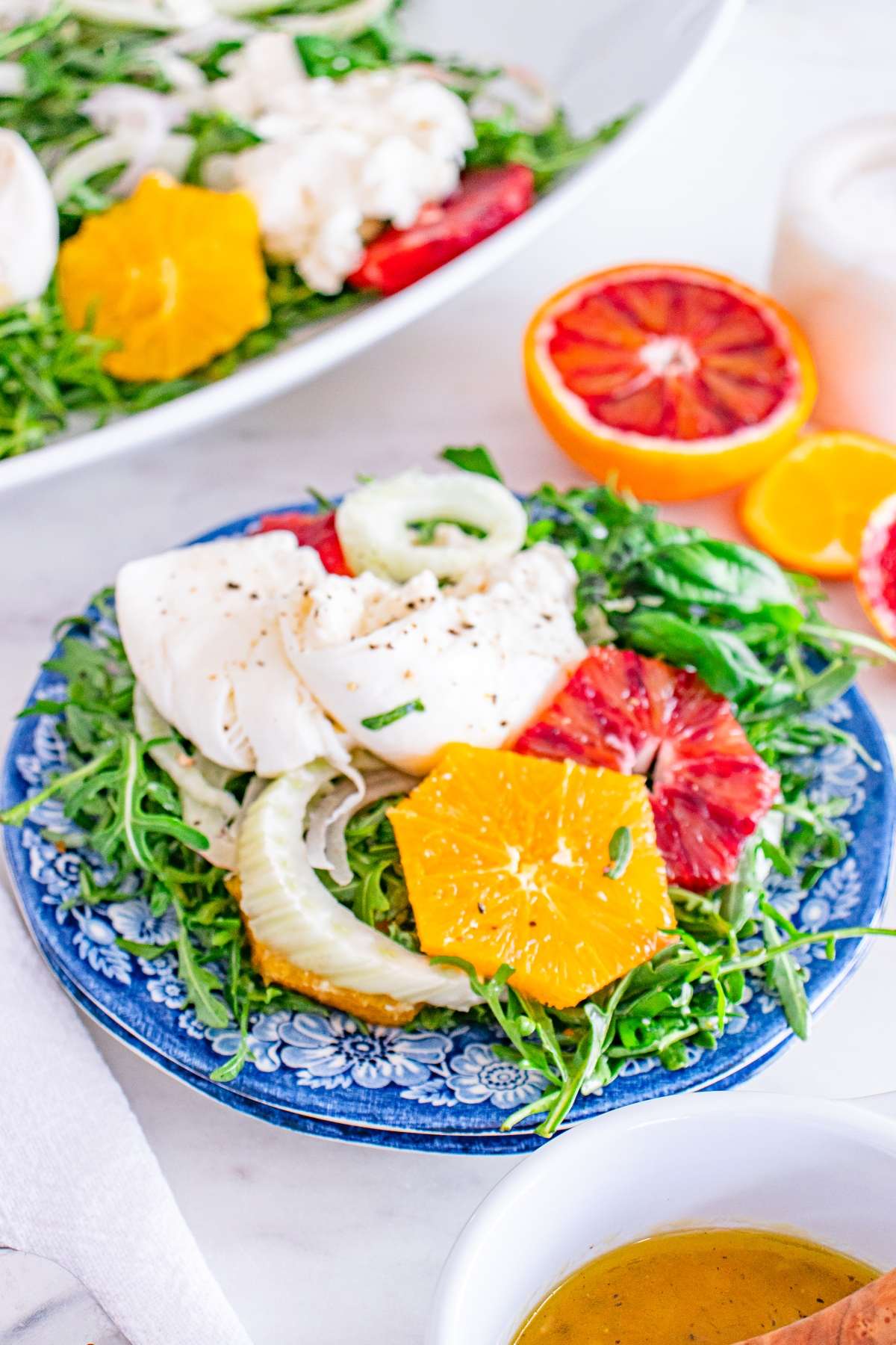 Colorful salad with arugula, orange slices, and cheese on a blue plate, next to an orange half and dressing.