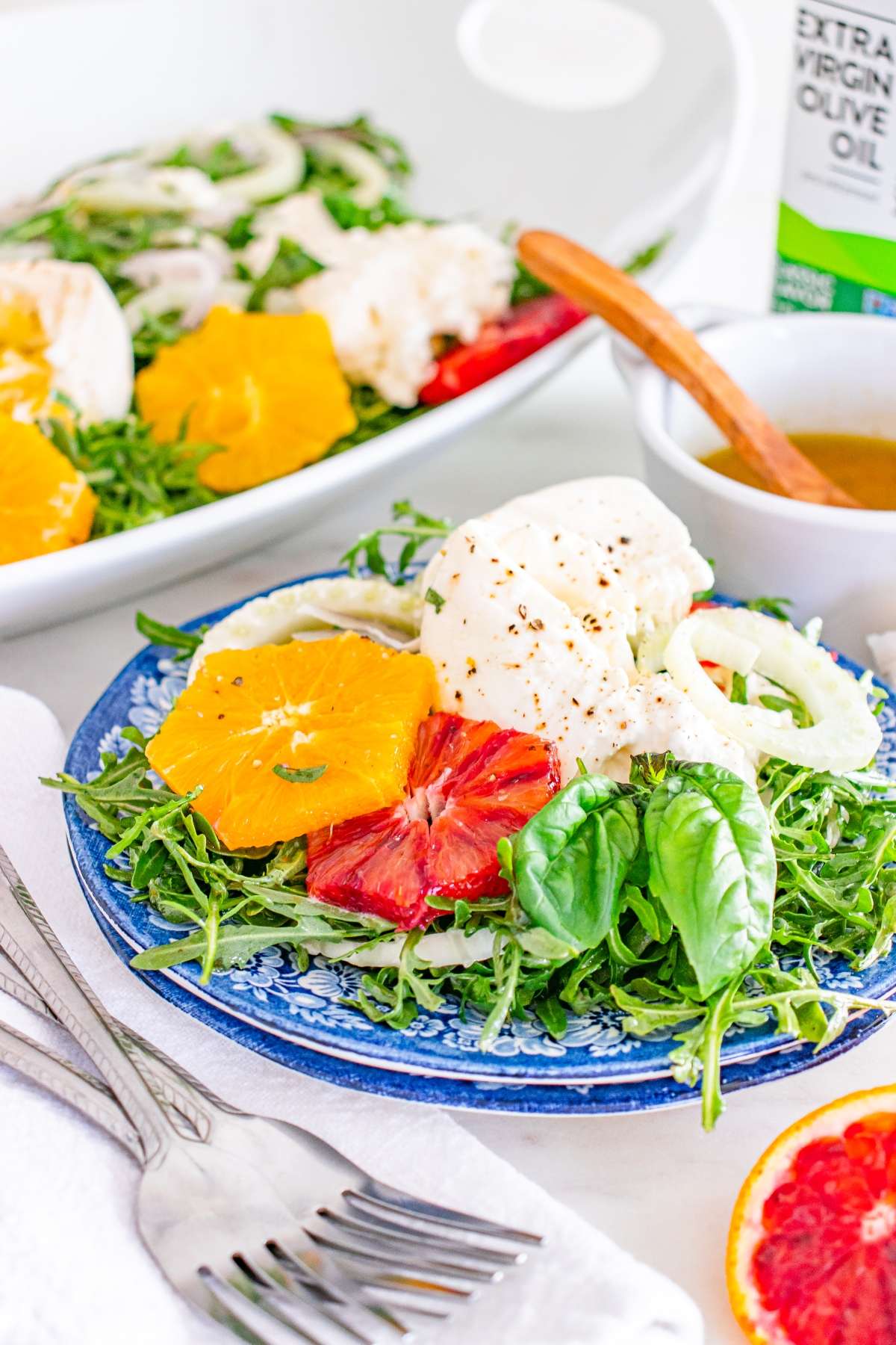 Plate with arugula, orange slices, burrata, and basil. Dressing and olive oil nearby. Fork on white napkin.