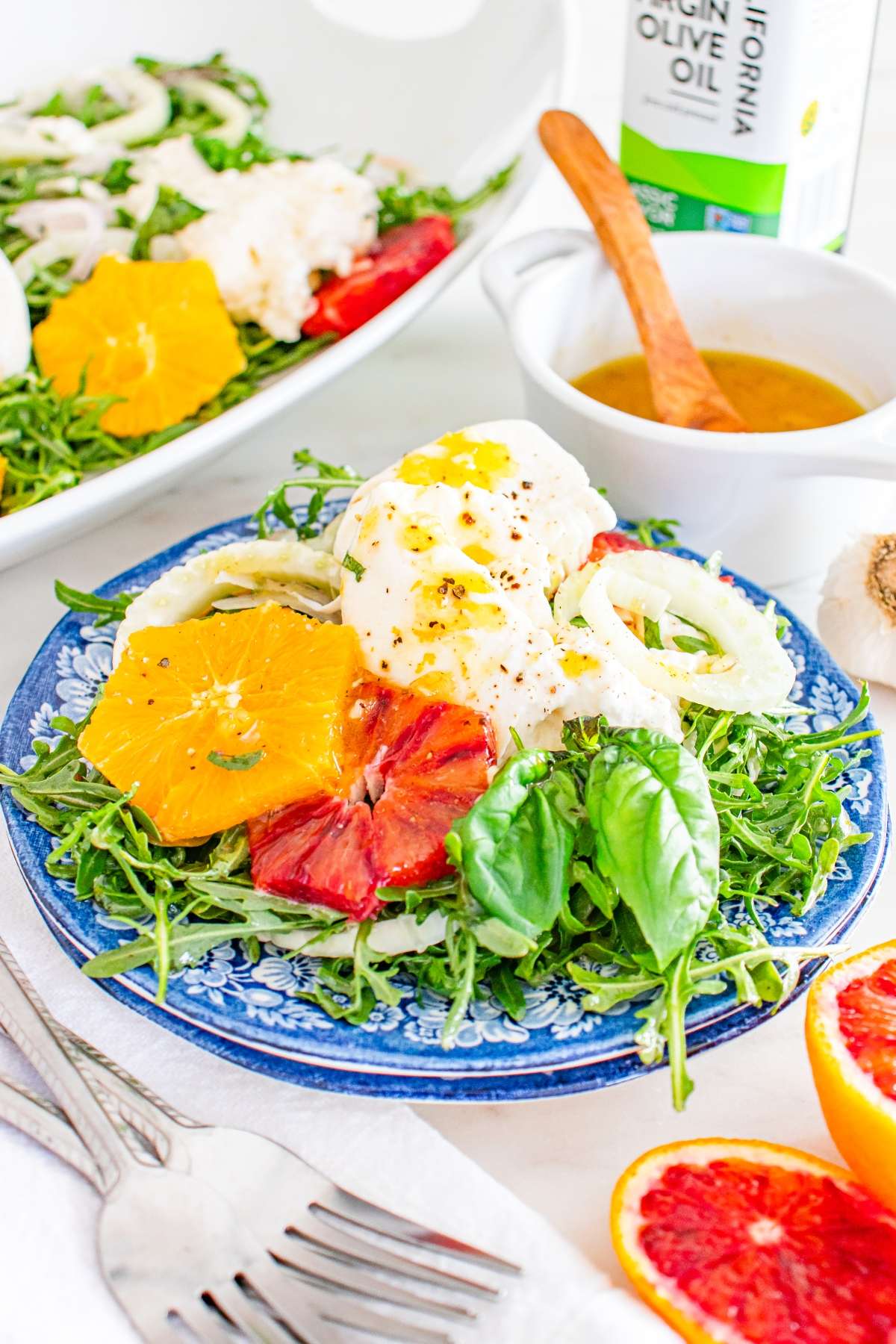 Salad with burrata, orange slices, arugula, and basil on a blue plate, with dressing and olive oil nearby.
