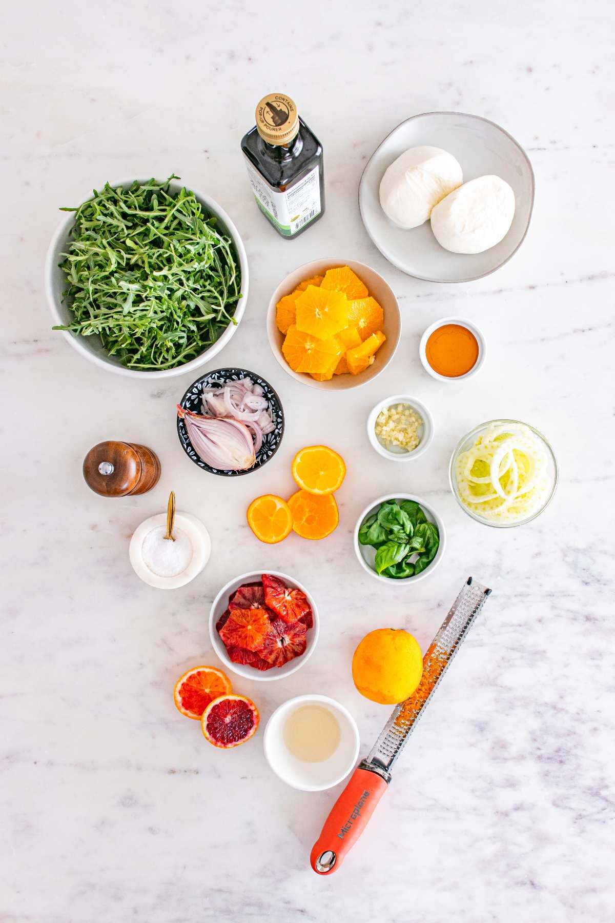 Ingredients for a salad including citrus, arugula, burrata, and seasonings arranged on a marble surface.