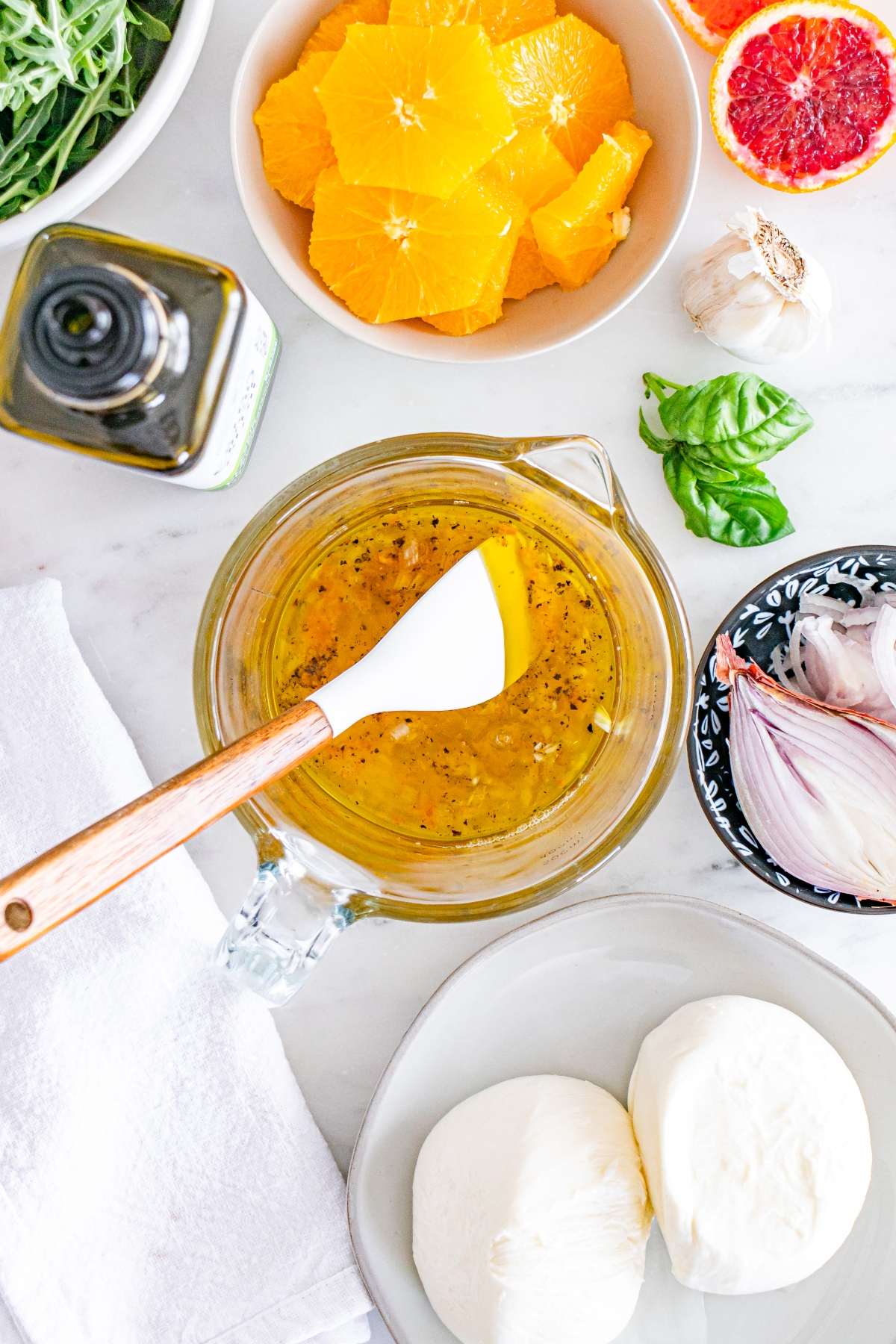 Ingredients for a salad on a marble surface: orange slices, mozzarella, basil, garlic, olive oil, and a jar with dressing.