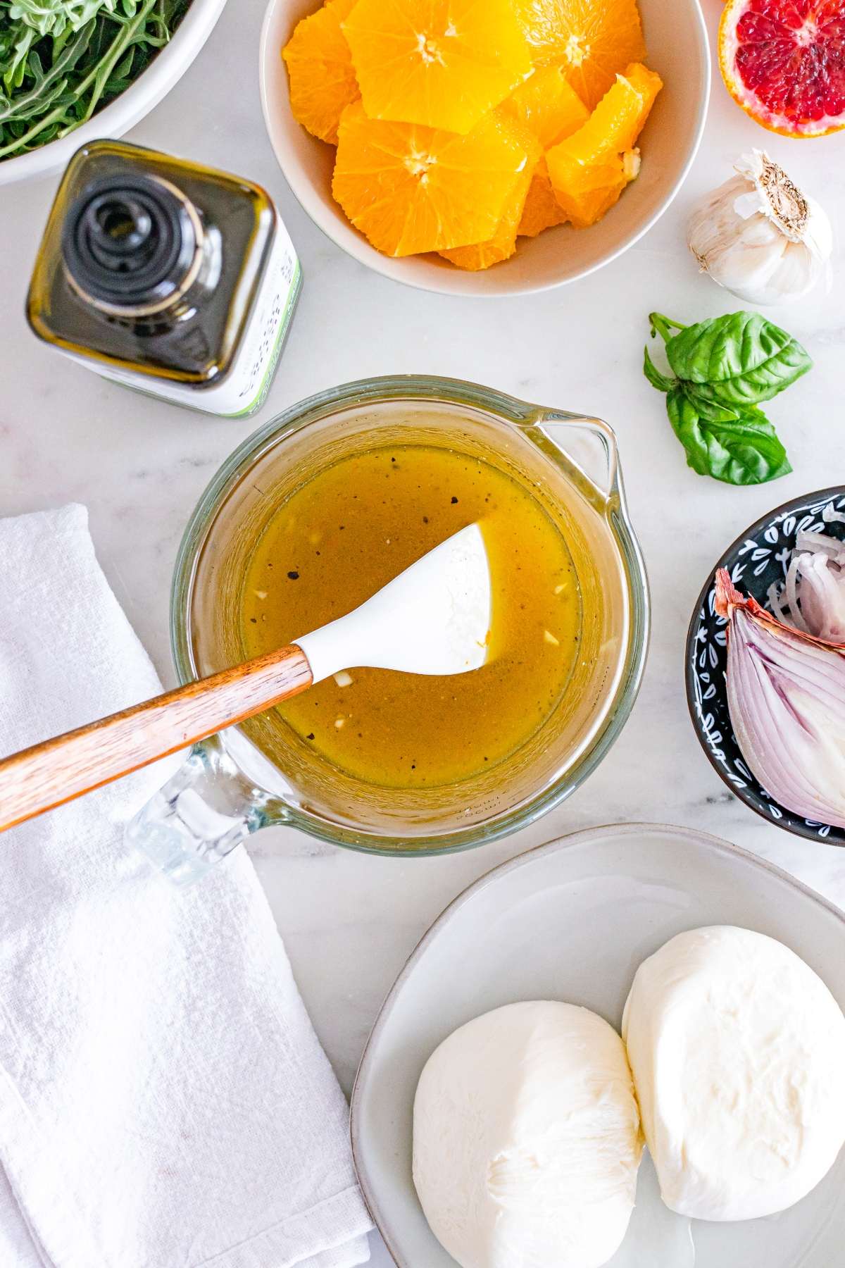 Bowl of citrus dressing with a spatula, surrounded by oranges, garlic, mozzarella, and shallots on a marble surface.