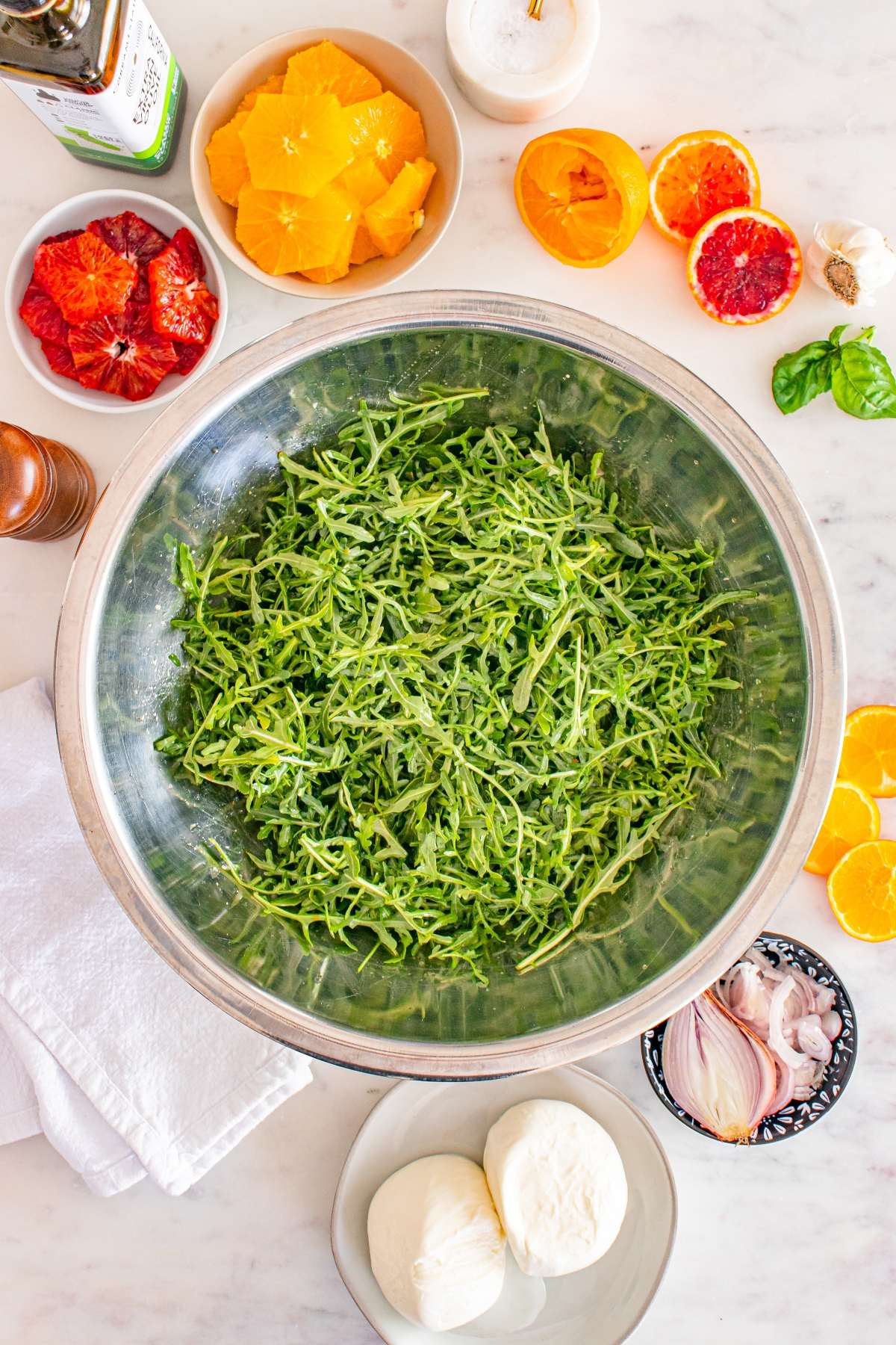 Fresh arugula in a bowl surrounded by citrus fruits, mozzarella, and seasonings on a marble countertop.