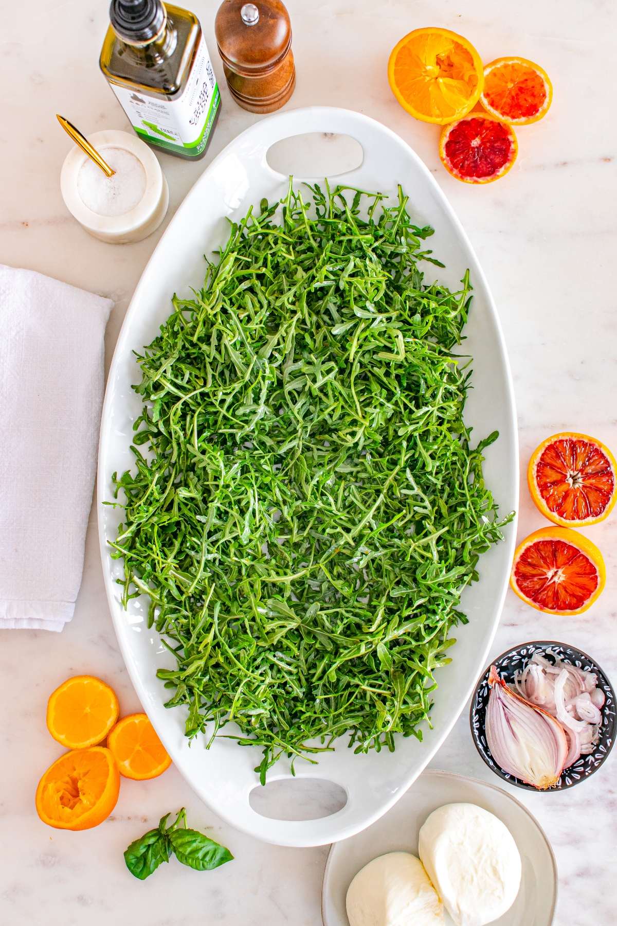 Oval platter of arugula on a table with orange slices, mozzarella, shallots, olive oil, salt, and pepper.