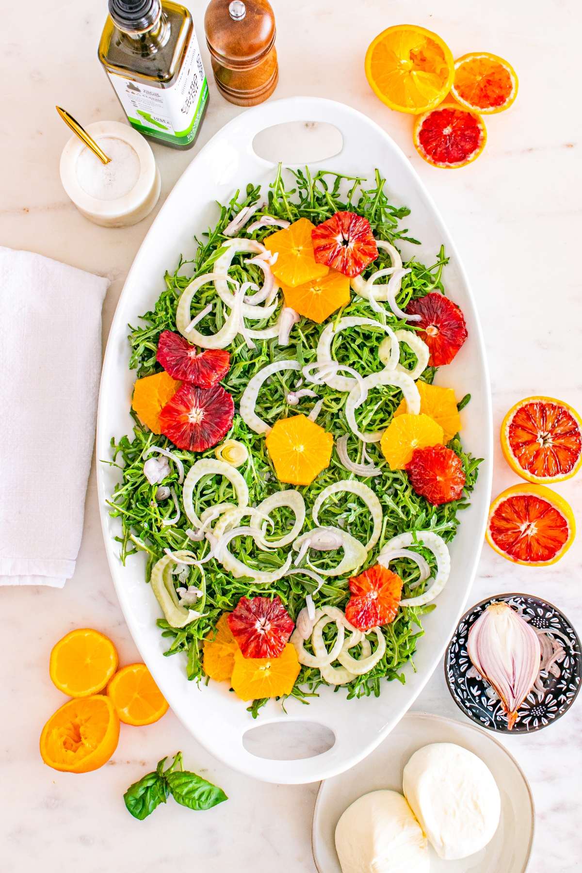 Oval platter with arugula, sliced fennel, and citrus fruits. Surrounding items include olive oil, spices, and mozzarella.