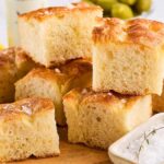 Sliced focaccia bread, crafted from a timeless recipe, is stacked on a wooden board, garnished with sea salt. Olives and oil linger invitingly in the background.