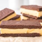 Homemade ice cream sandwiches with chocolate cookies and creamy vanilla filling rest on parchment paper, with more sandwiches tempting in the background.