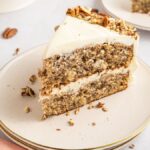 Slice of layered cream cake with pecans, served on a white plate, accompanied by a peach napkin and forks.