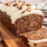 A delightful hummingbird loaf cake with cream cheese frosting and pecans rests on a wooden board, accompanied by a knife and napkin, ready to be savored.