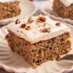 A slice of frosted hummingbird sheet cake with nuts, rests on a scalloped plate, accompanied by a fork.