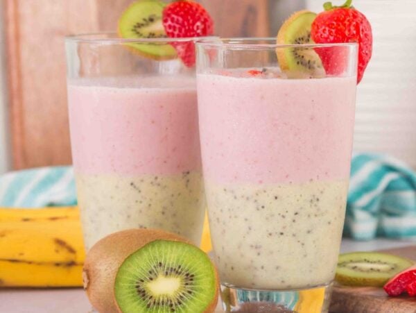 Two Layered Kiwi Strawberry Smoothie glasses, topped with fresh strawberry and kiwi slices, with bananas and kiwis in the background.