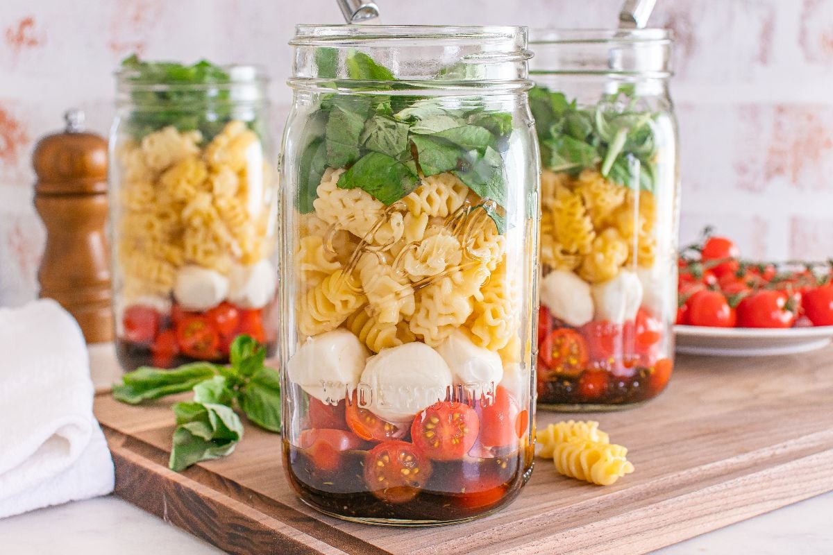 Three mason jars filled with Caprese pasta salad—layered pasta, cherry tomatoes, mozzarella balls, and fresh basil—sit on a wooden board.
