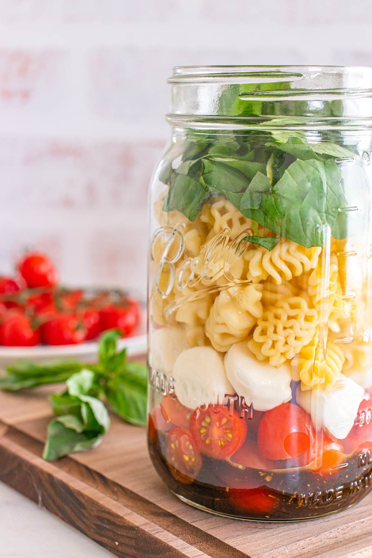 A mason jar layered with pasta, mozzarella, cherry tomatoes, basil, and dressing, on a wooden board.