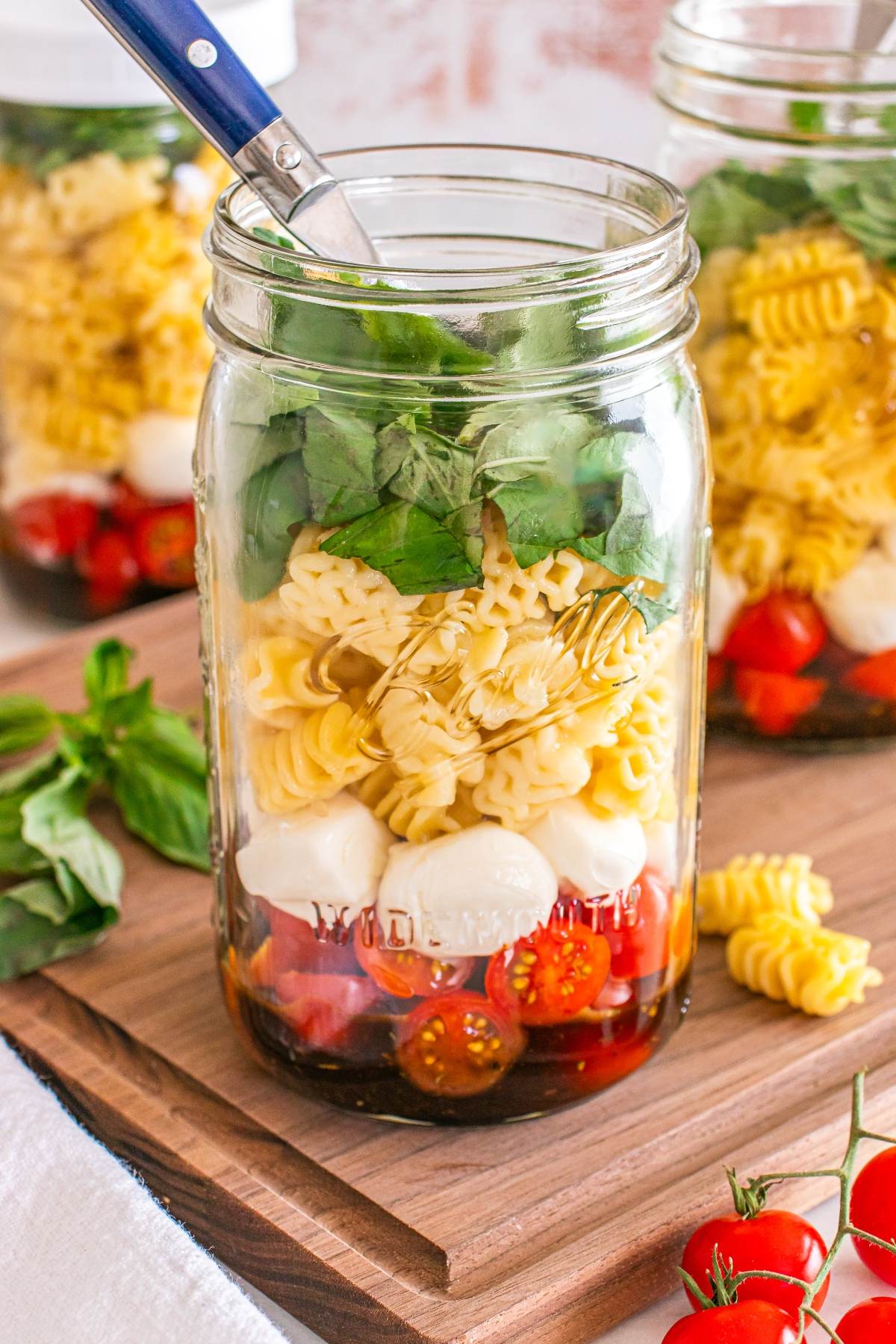 A mason jar filled with pasta, cherry tomatoes, mozzarella balls, and fresh basil, with a fork inside.