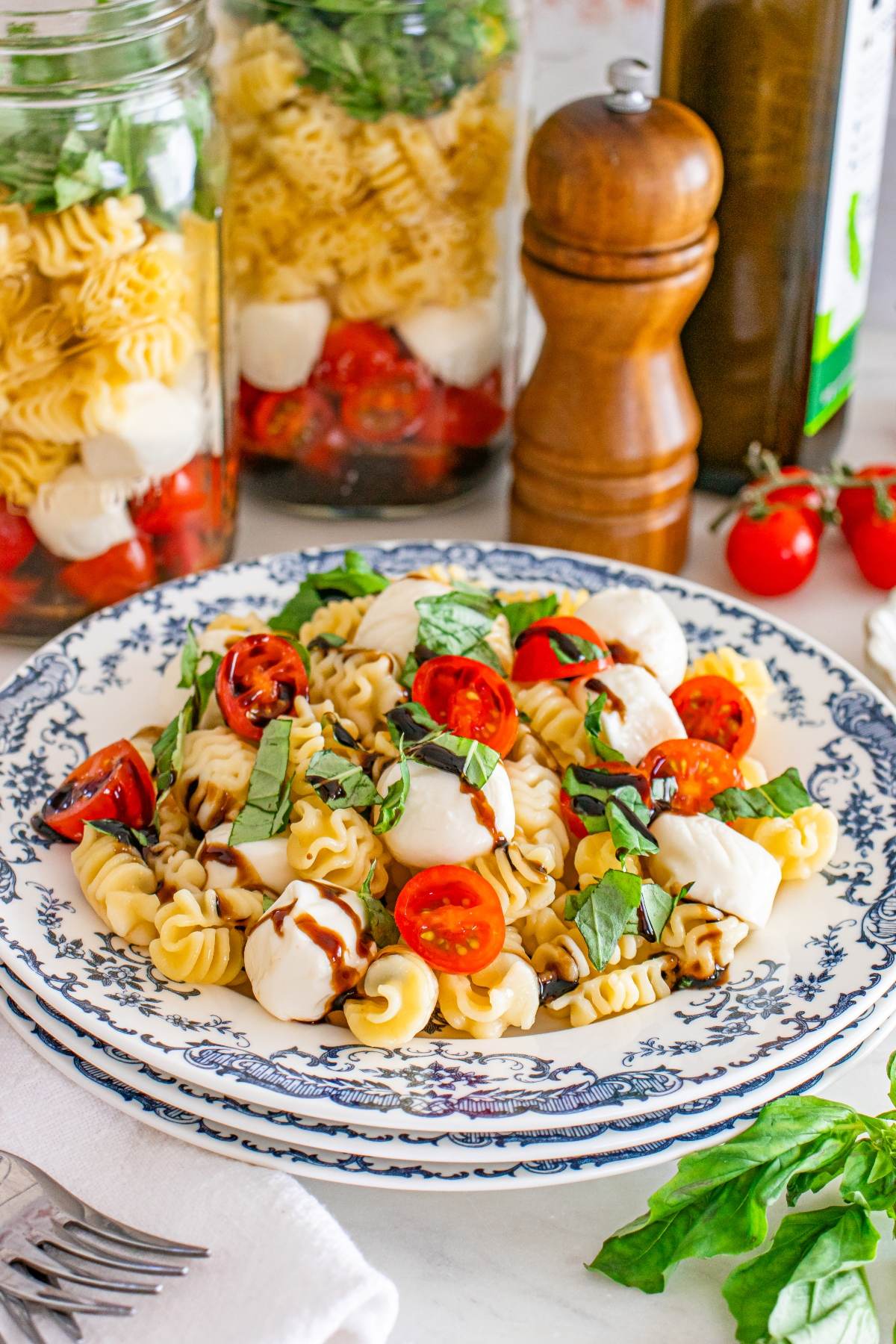 Pasta salad with cherry tomatoes, mozzarella, and basil on a blue floral plate, with jars and pepper mill behind.
