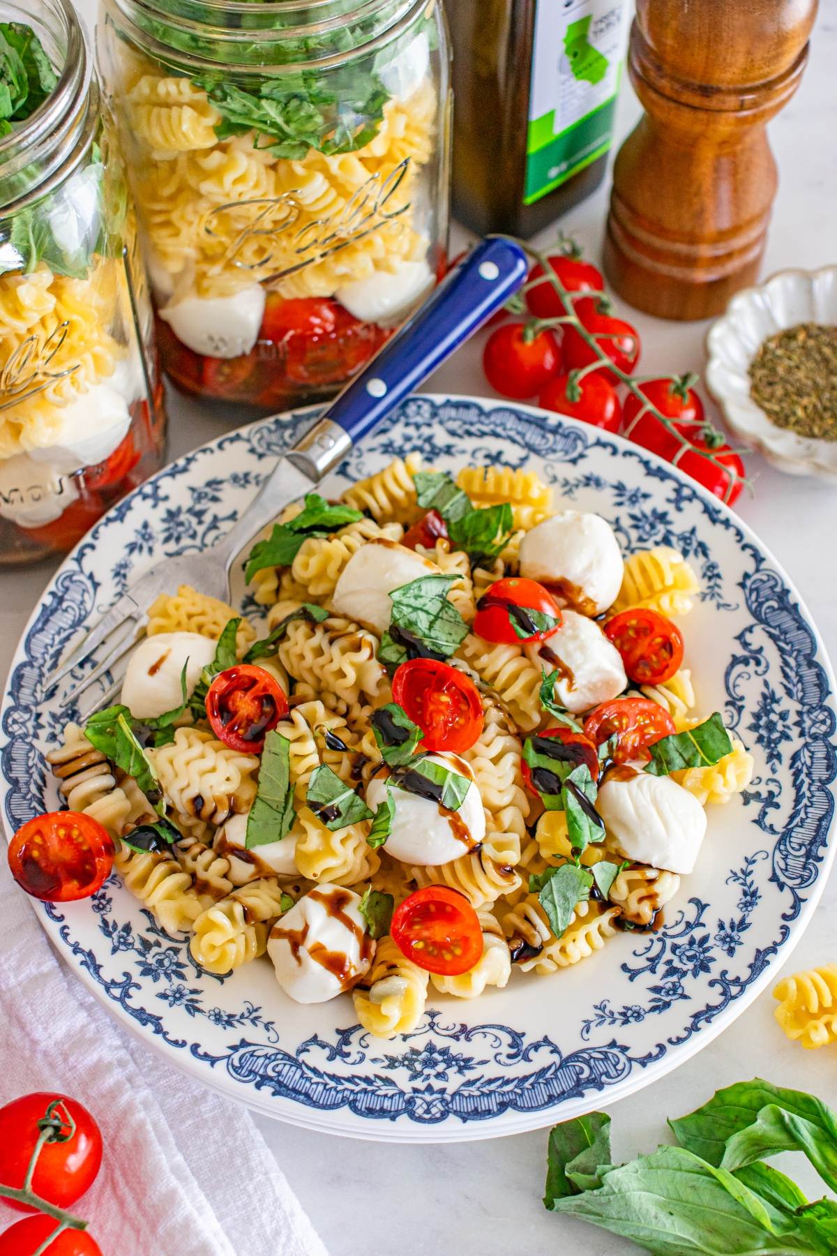 Pasta salad with tomatoes, mozzarella, basil, and dressing on a blue floral plate, with jars of pasta nearby.