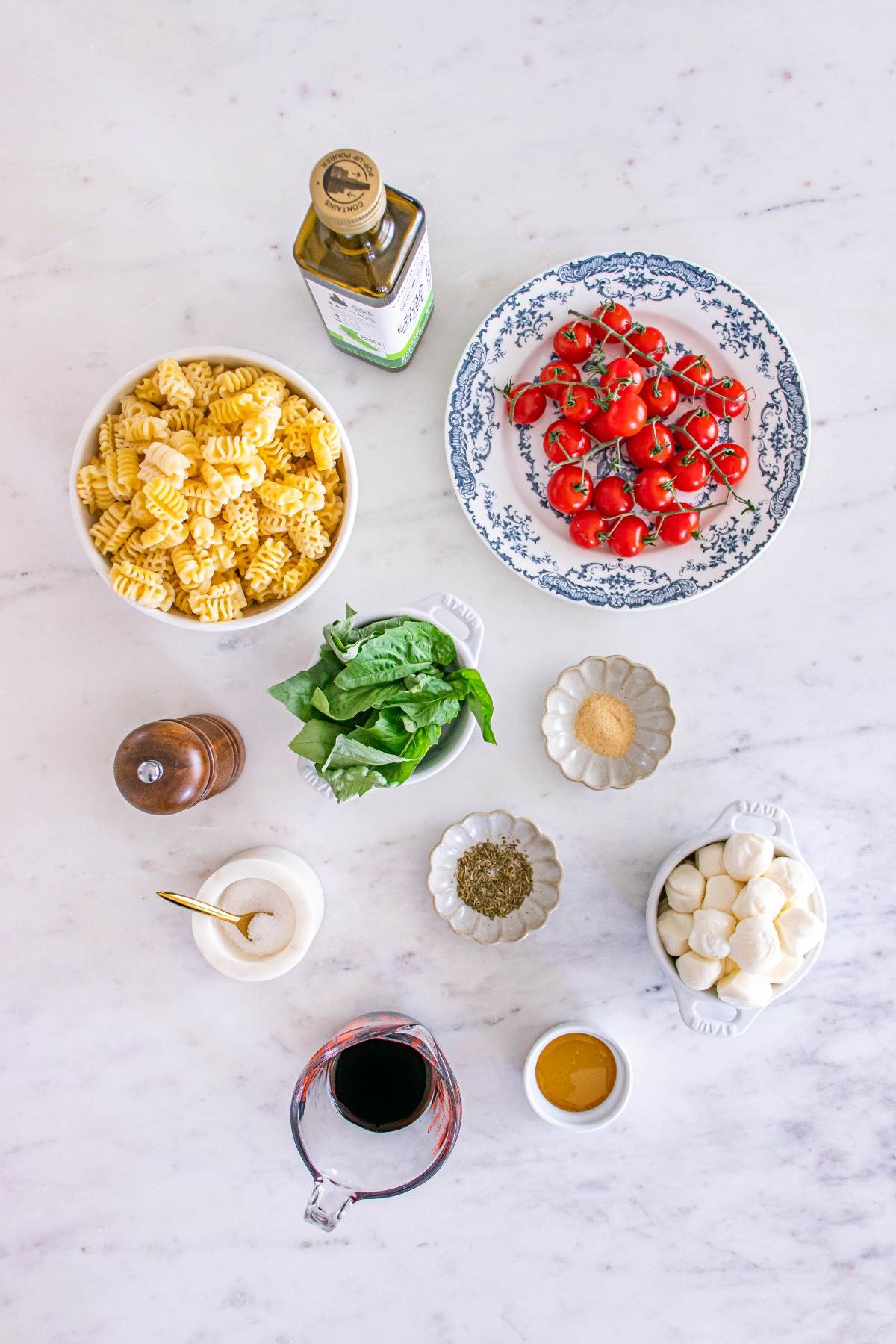Pasta, tomatoes, mozzarella, basil, olive oil, vinegar, spices, and honey are arranged on a white countertop.