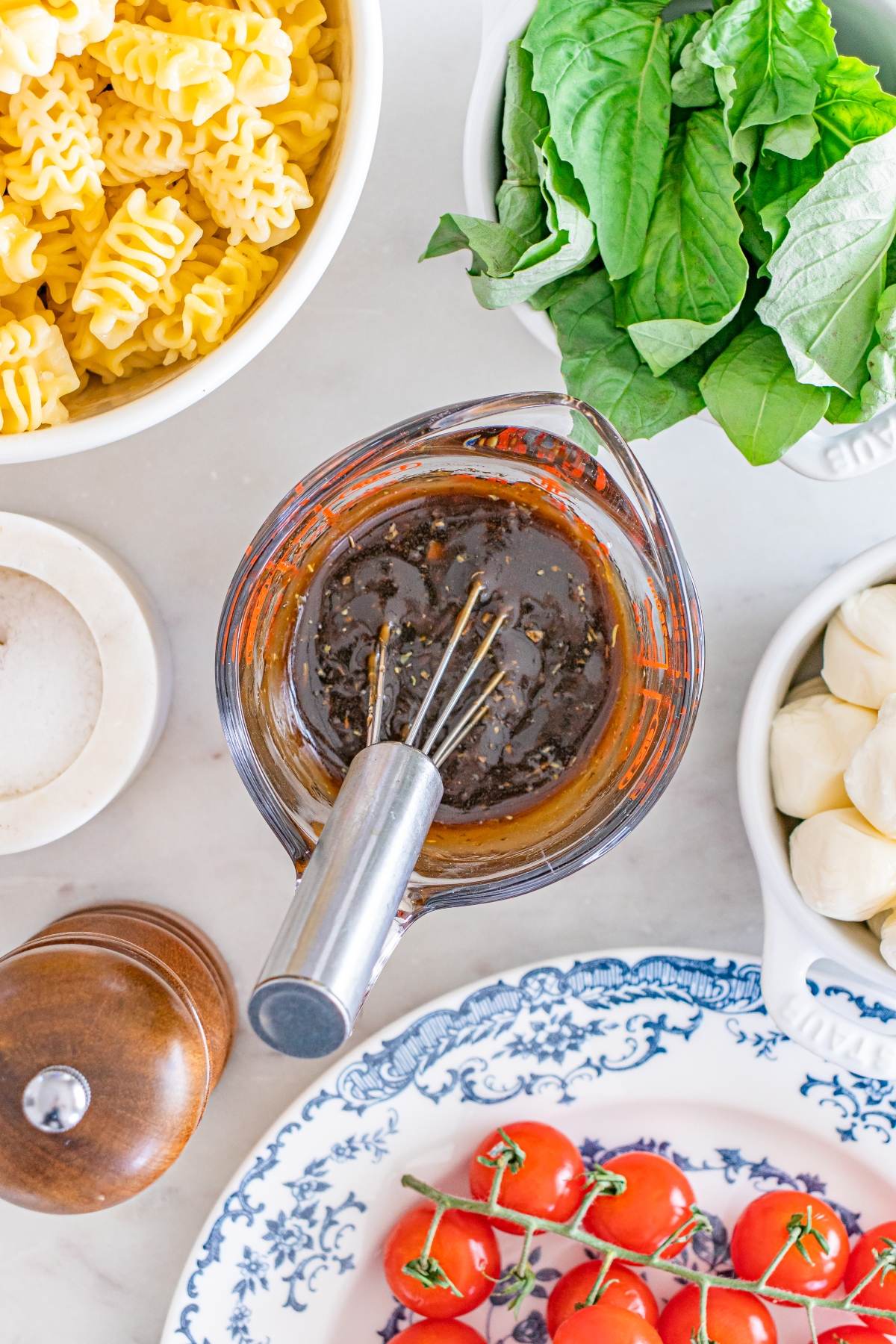 A bowl of dark vinaigrette with a whisk, surrounded by pasta, basil, mozzarella, tomatoes, salt, and pepper.