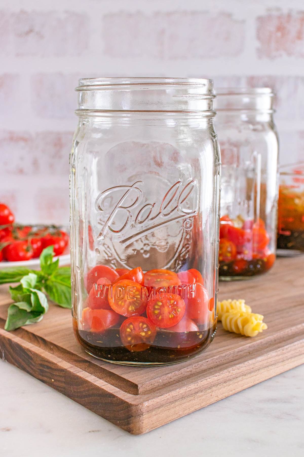 A mason jar with sliced cherry tomatoes inside, sitting on a wooden board with pasta and basil nearby.