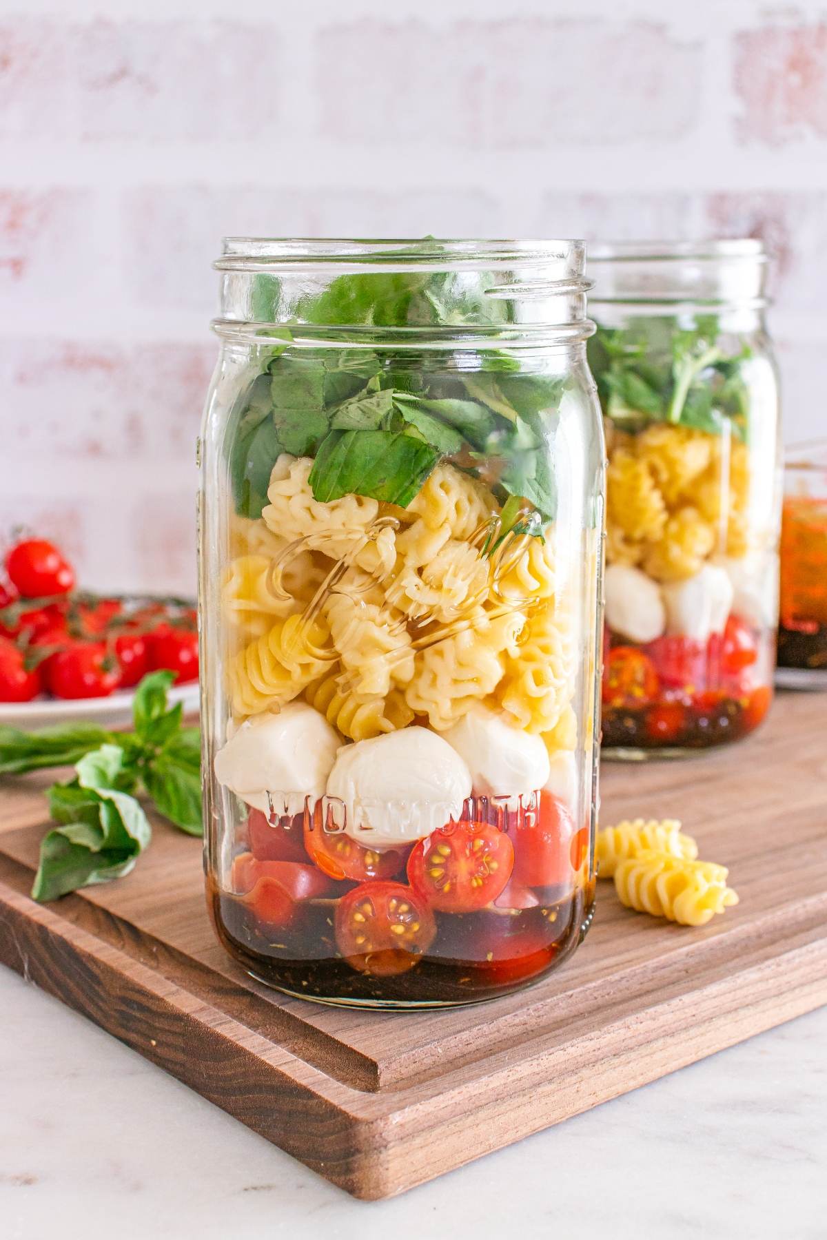 Mason jars filled with pasta, cherry tomatoes, mozzarella balls, spinach, and basil on a wooden board.