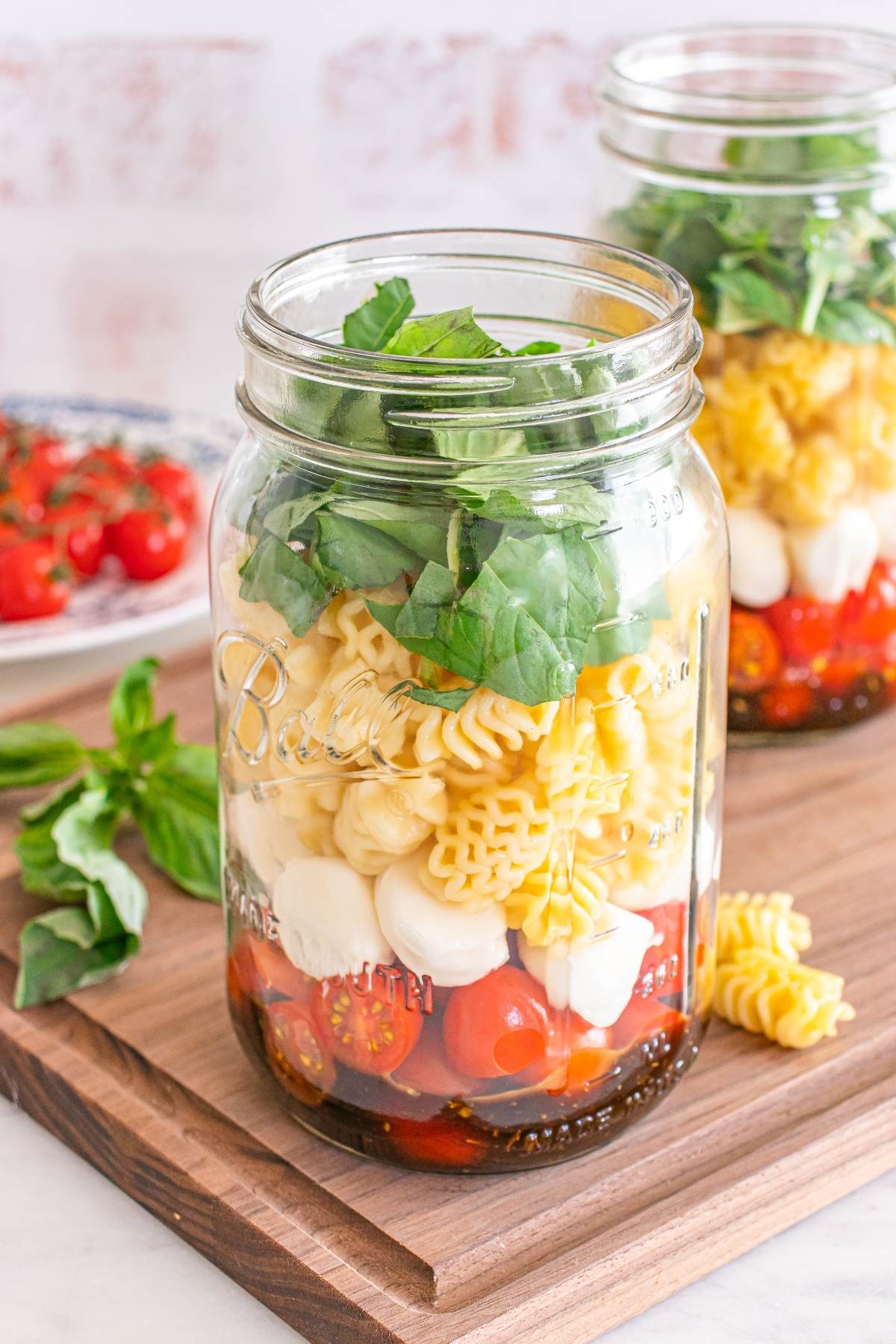 A mason jar filled with pasta, cherry tomatoes, mozzarella balls, and fresh basil on a wooden board.