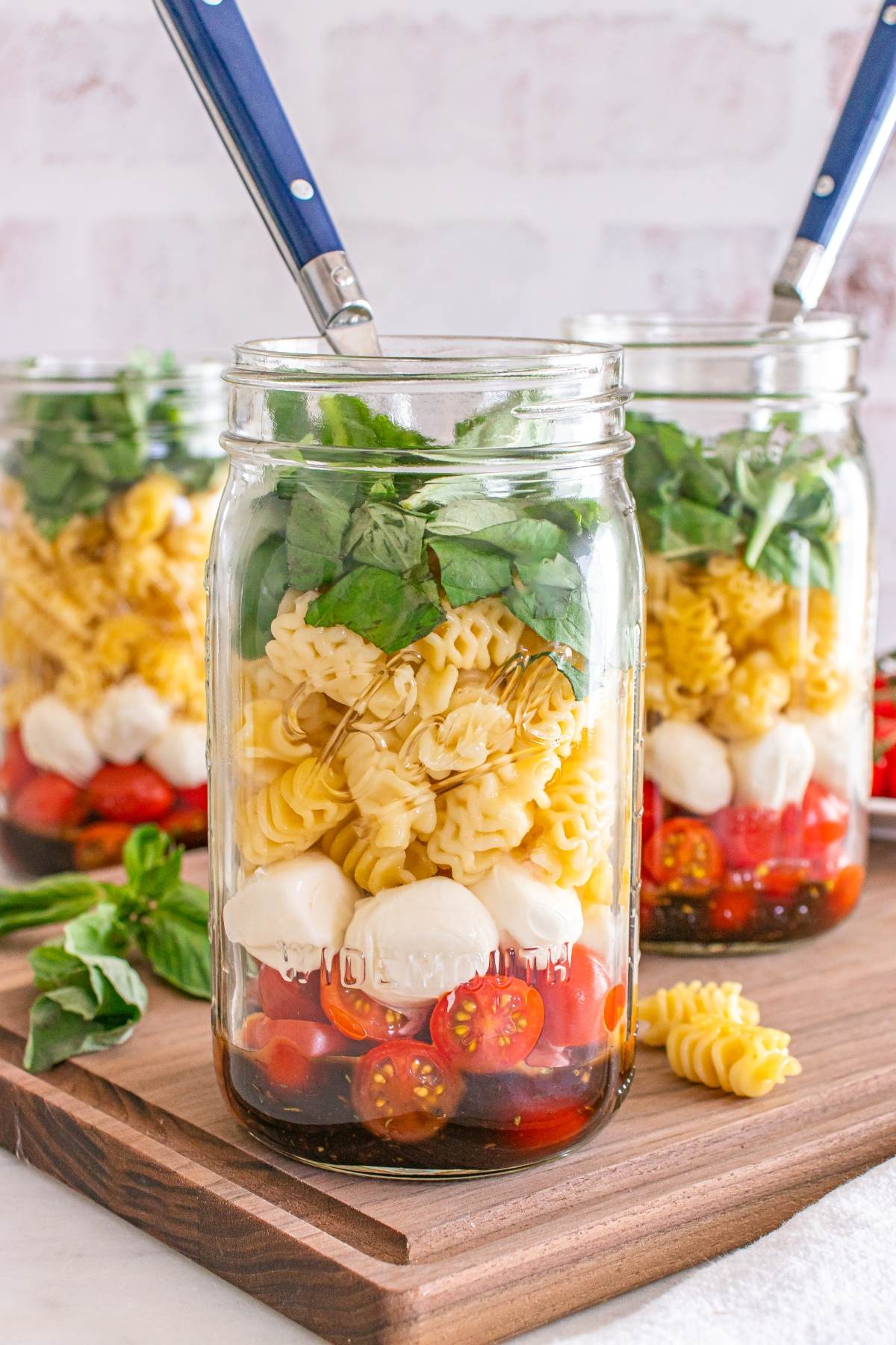 Three mason jars filled with pasta, cherry tomatoes, mozzarella, spinach, and utensils on a wooden board.