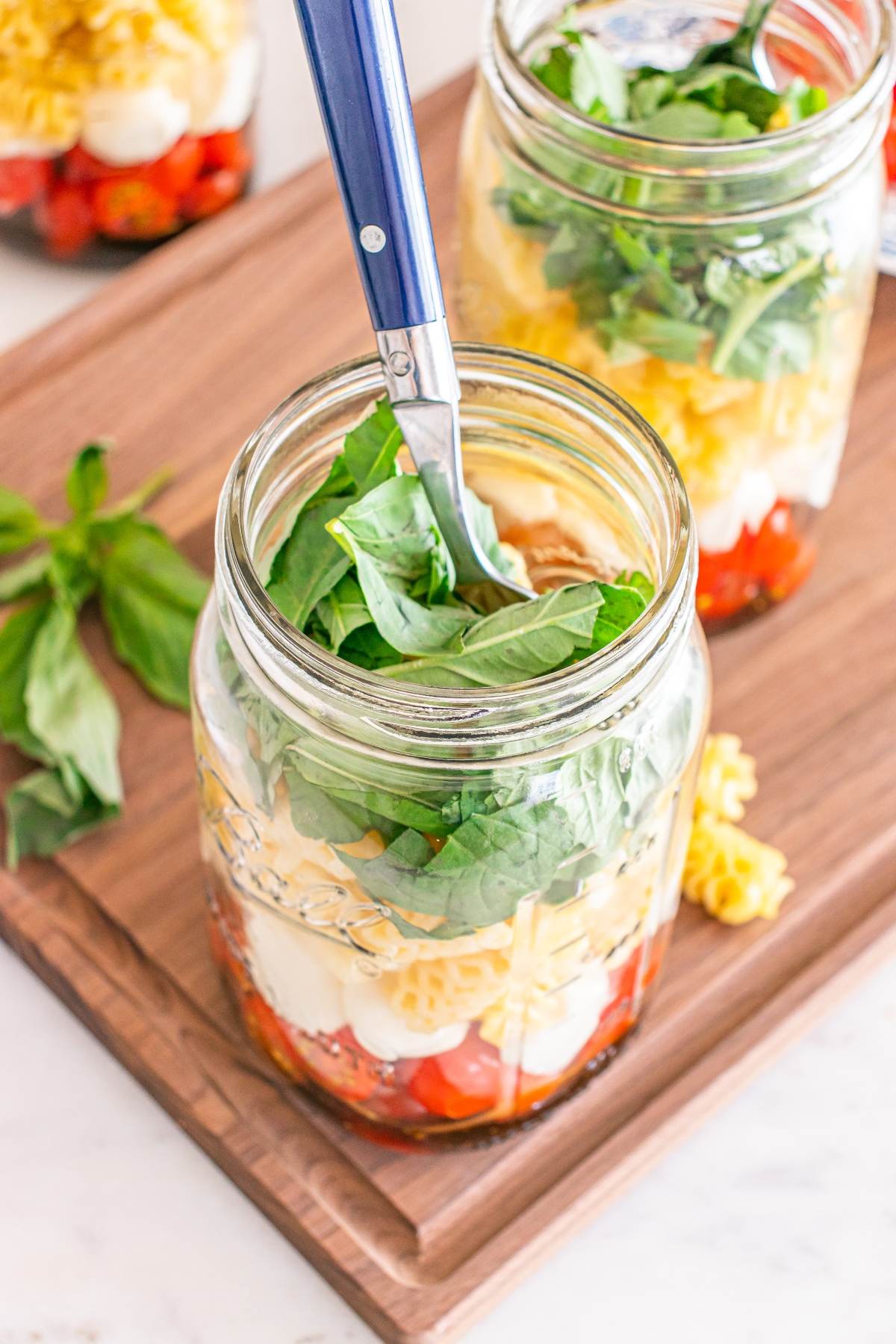 A mason jar filled with pasta, tomatoes, mozzarella, and fresh basil, with a blue-handled fork inside.