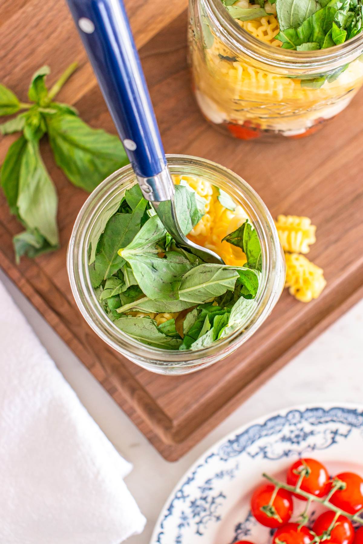 A mason jar filled with pasta, fresh basil, and vegetables sits on a wooden board with a fork inside.