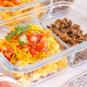 Glass meal prep container with scrambled eggs, ground meat, shredded cheese, and diced tomatoes in separate sections—perfect for assembling easy meal prep breakfast taco bowls.