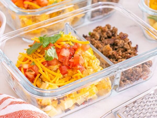 Glass meal prep container with scrambled eggs, ground meat, shredded cheese, and diced tomatoes in separate sections—perfect for assembling easy meal prep breakfast taco bowls.