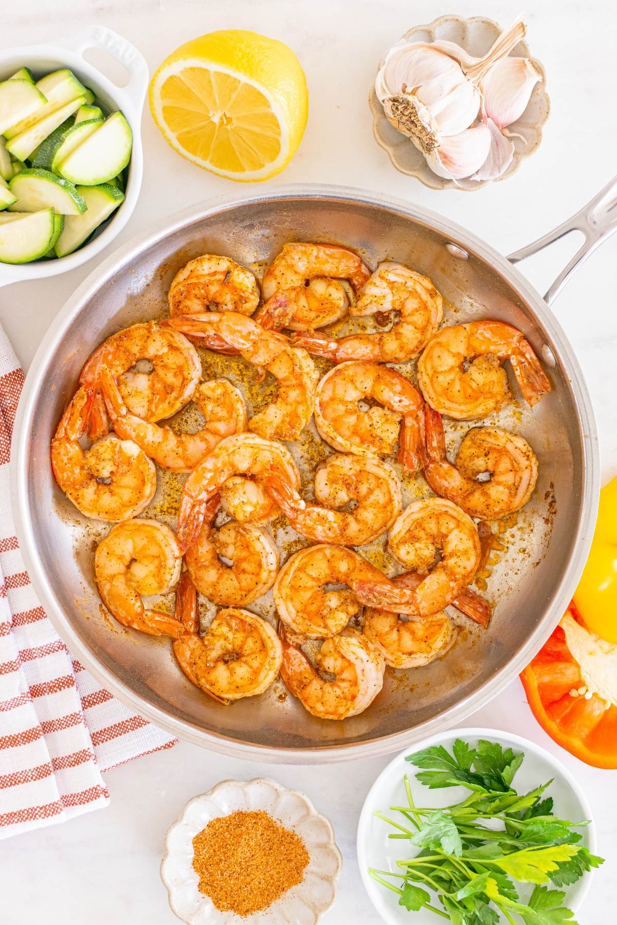 Shrimp cooking in a skillet, surrounded by fresh vegetables, lemon, garlic, and spices on a white surface.