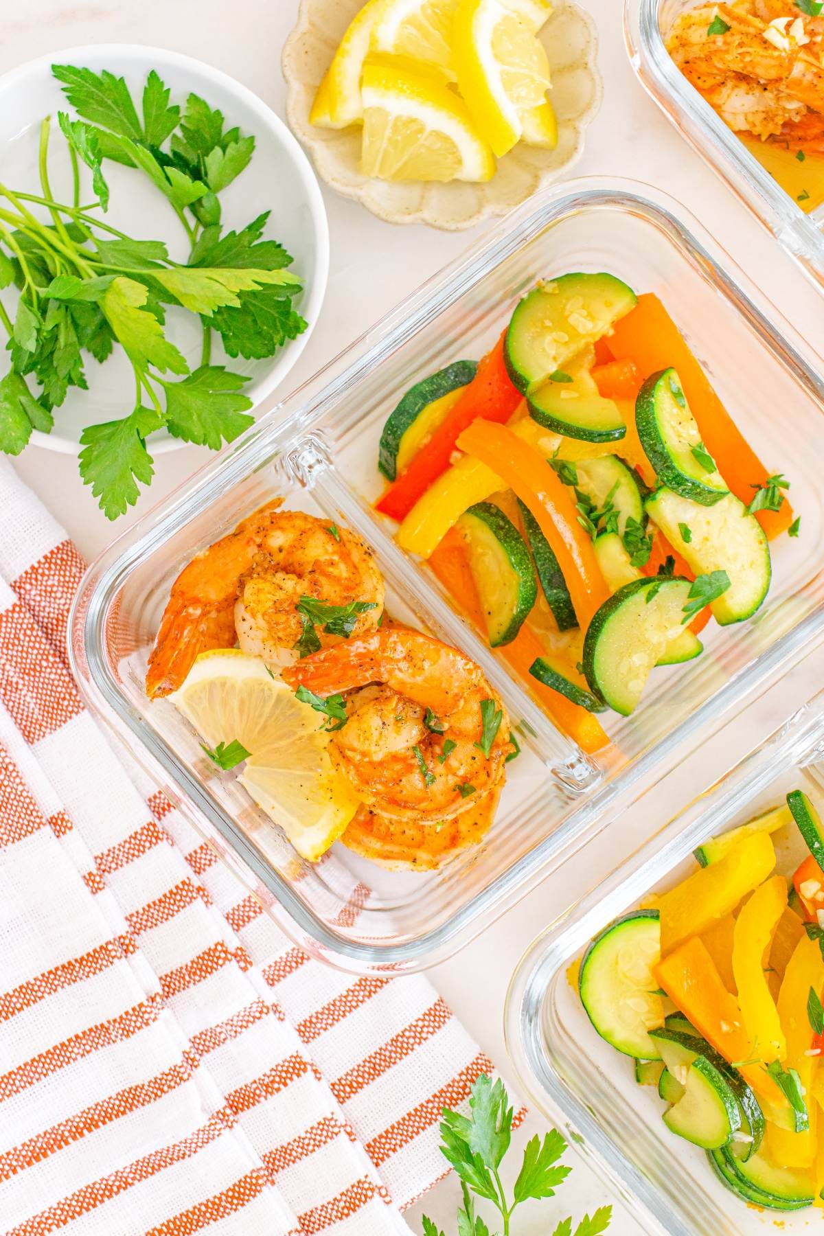 Glass containers with shrimp, lemon slices, bell peppers, and zucchini, next to parsley and lemon wedges.