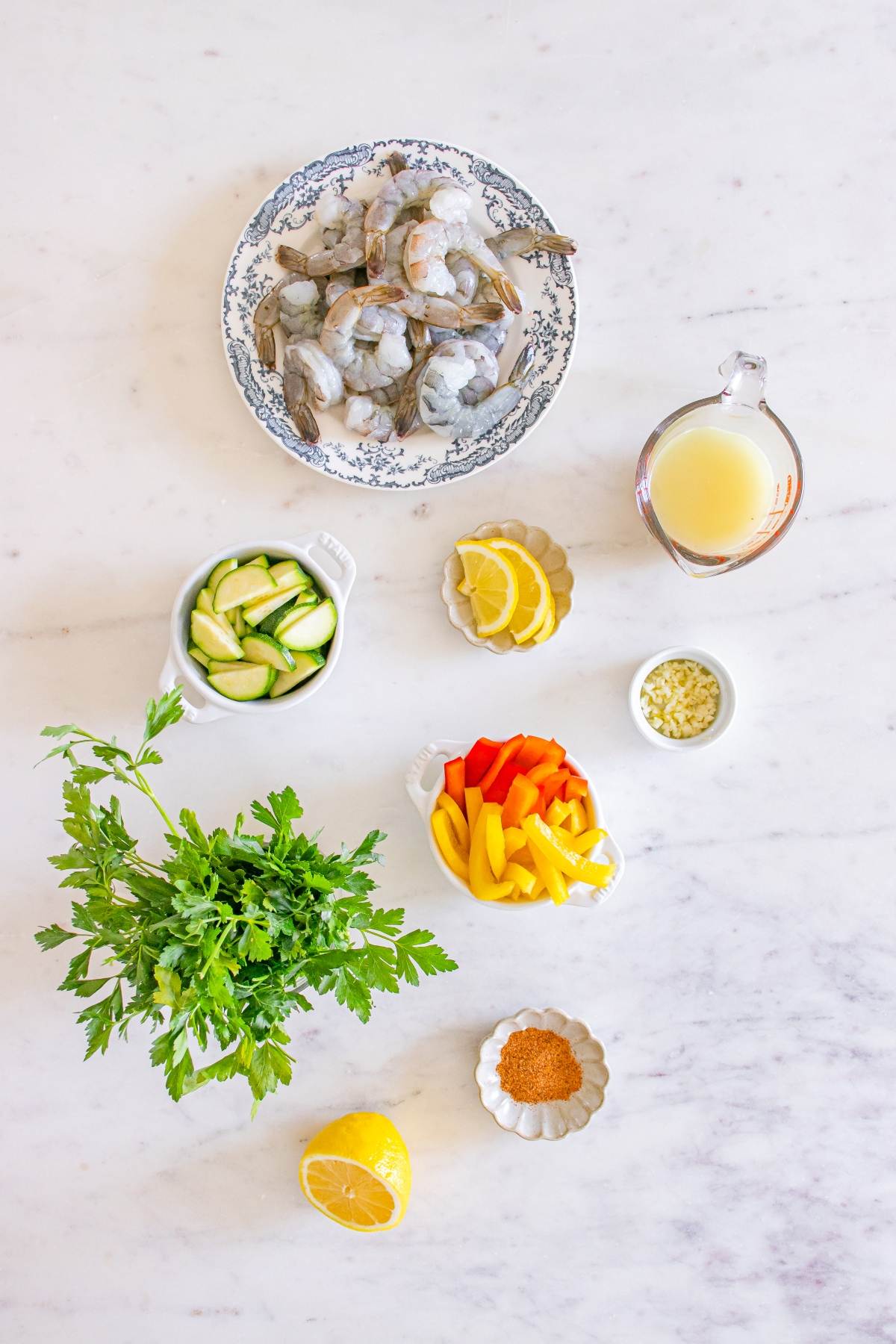 Shrimp, sliced zucchini, bell peppers, parsley, lemon, broth, spices, and garlic on a white marble surface.
